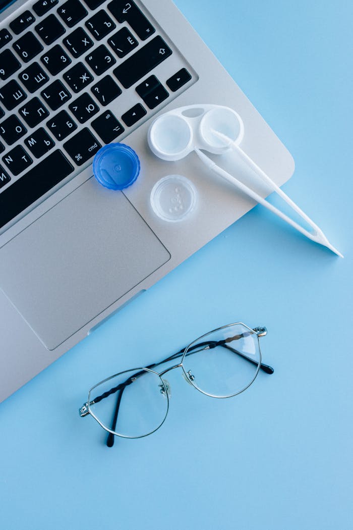 gallery-5 Flat lay of eyeglasses, contact lens case, and laptop on a blue surface.